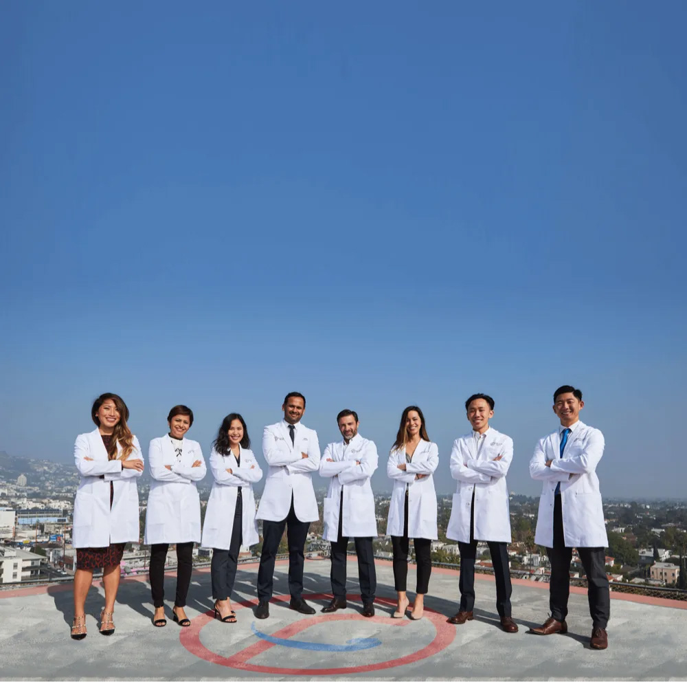 A group of seven medical professionals posing together outdoors with a clear blue sky background.