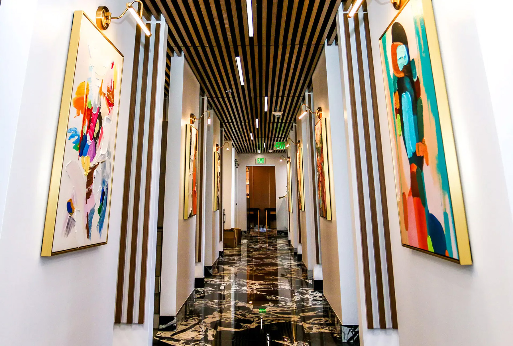 The image shows an interior hallway with modern decor, featuring artwork displayed on the walls, bright lighting, and a series of paintings aligned along the corridor.