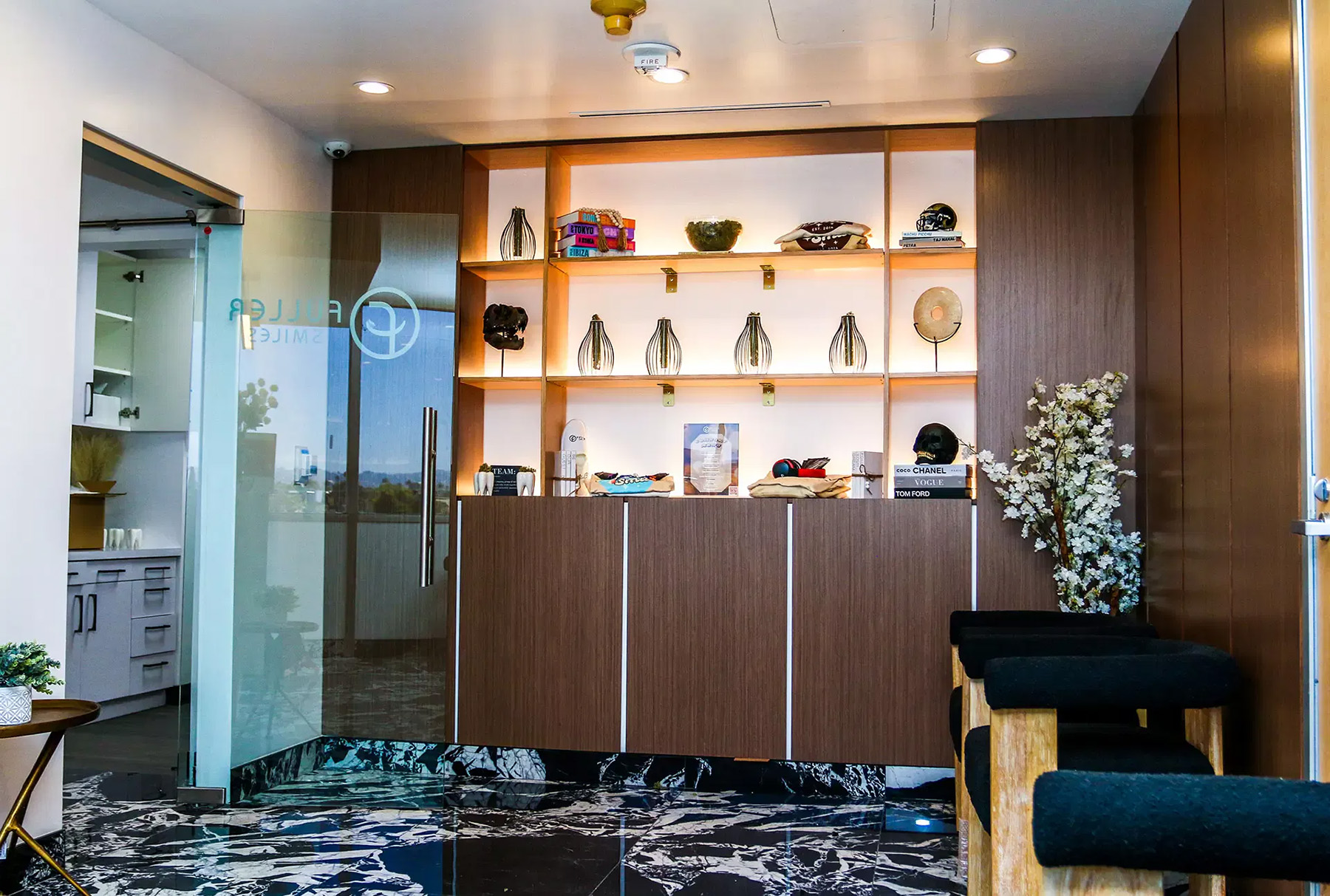 The image shows an interior space with modern decor, featuring a wooden cabinet displaying various items, a glass wall with a dark metal frame, and a reception area with seating and a countertop.